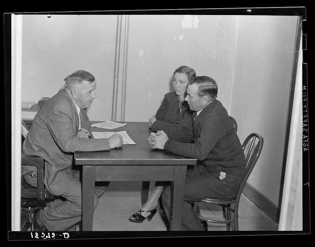 The image is a black and white photograph depicting three individuals seated around a table engaged in what appears to be an interview or discussion. The setting suggests it could be from the early-to-mid 20th century, given the style of clothing worn by the subjects.

On one side of the table sits an older man with gray hair, wearing glasses and dressed in a dark suit jacket over a lighter shirt paired with trousers. His posture is relaxed as he looks towards his interlocutor on the opposite side.

Across from him are two younger individuals: a woman seated slightly behind a man who appears to be listening intently or perhaps asking questions. The woman has her hands clasped and gazes directly at the older gentleman, while the second individual sits with one leg crossed over the other knee, sporting a dark suit similar in style but lighter shade than that of his counterpart.

The table holds some papers which could indicate documents relevant to their discussion. In the background is a plain wall with minimal decoration or furnishings visible; this stark setting suggests an office environment focused on serious matters like work, meetings, or interviews during that historical period.