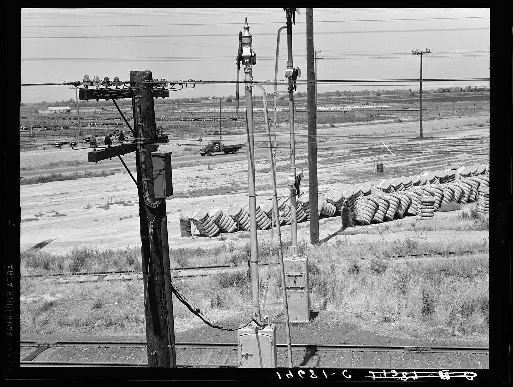 This black-and-white photograph depicts an industrial or agricultural landscape, likely from the mid-20th century given its style. In the foreground are utility poles and electrical wires running parallel to a railway track on which no train is visible. The background reveals stacks of burlap sacks or bags in neat rows across what appears to be farmland or waste land with sparse vegetation.

A notable feature includes an old-fashioned pickup truck, suggesting transportation activity related to the nearby railroad tracks. In the distance, one can discern a small building and more utility poles, indicating infrastructure for either industrial purposes or agricultural activities.

The photograph is labeled "1028- C" at its bottom right corner, possibly referencing a cataloging system used by the photographer Dorothea Lange during her time in California capturing scenes of people affected by social issues. The photo's composition and subject matter suggest it may be from an era where manual labor or rural agricultural practices were prominent.

Further context indicates this could have been taken between Tulare and Fresno, California, based on information provided with the image link to a source detailing Lange’s work in that region during her lifetime, particularly focusing on socio-economic conditions.