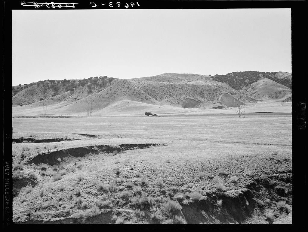 The photo is a black-and-white landscape depicting a vast expanse of flat, barren land with sparse vegetation. In the distance, rolling hills rise up against an overcast sky. Dominating one side of the hilltops are electric transmission towers and lines stretching into the horizon. A single vehicle appears in the far background near these structures, indicating human presence or activity. The ground is littered with small shrubs and dry grasses that suggest a semi-arid climate, possibly indicative of a drought-affected region. There's no visible vegetation suggesting lush greenery typically associated with well-watered environments like forests or fields. A watermark on the right side indicates it was taken by Dorothea Lange.
