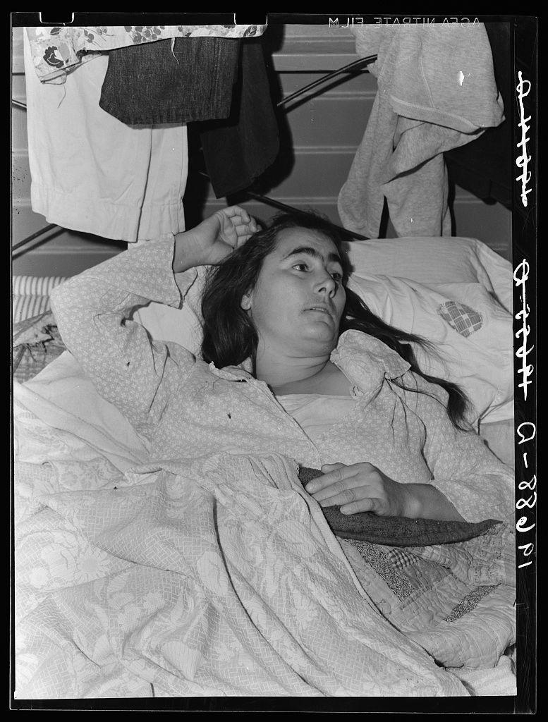 The image is a black and white photograph showing an individual lying down, seemingly resting or sleeping. The person appears to be wearing pajamas with a patterned design on the top garment, possibly featuring hearts or similar shapes. They have dark hair that falls over their shoulders and are covered up to just below the chest area by what seems like a blanket or bedspread.
In the background, there is another individual sitting upright in a chair partially obscured from view, wearing casual clothing with visible denim pants and a light-colored top. The setting appears to be an indoor environment, possibly within a camp as indicated by various articles of clothes hanging on lines above. A pillow with a circular design or logo can also be seen.
The photo bears some textual markings: "Photo by Dorothea Lange" in the upper right corner and what seems like handwritten text at the bottom that is partially obscured but includes numbers, potentially indicating a date or other identification information related to the photograph's context. This image likely has historical significance as it appears to be part of an archive documenting migratory agricultural workers during their time spent in camps managed by the Farm Security Administration (FSA).