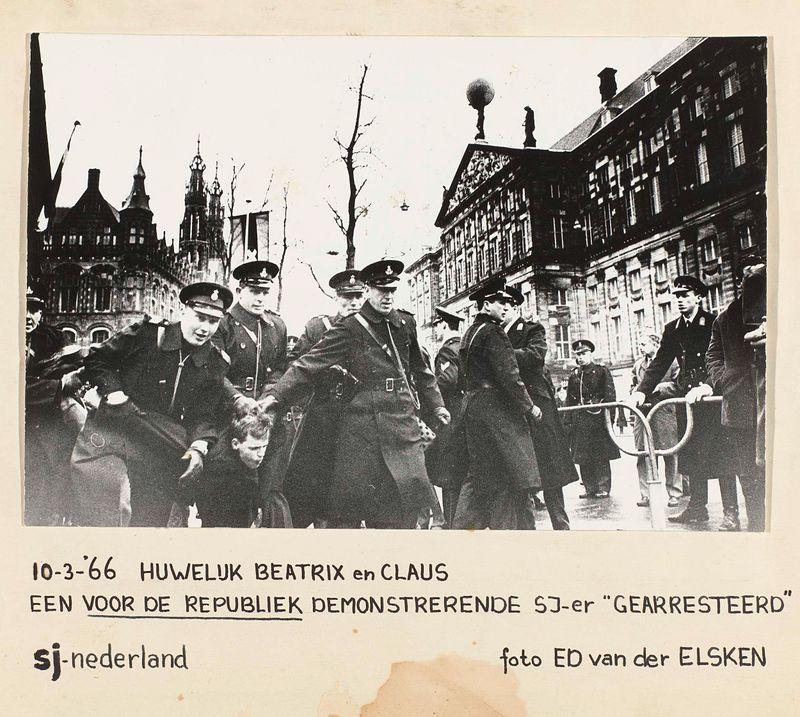 The image is a black and white photograph that depicts a group of people, predominantly dressed in dark military-style uniforms with peaked caps. They appear to be gathered outside on an urban street during the daytime, as indicated by natural light and shadows visible around them.

In the background are tall buildings with architectural features such as spires, chimneys, large windows, and ornate detailing that suggest a European cityscape, possibly in the Netherlands given the text present. The architecture hints at a historic or government district of Amsterdam due to recognizable structures like those found on Dam Square during the early 1960s.

The foreground shows several individuals close-up; one prominent figure is walking forward with determined strides and looking off-camera while others seem engaged in conversation among themselves, creating an impression of camaraderie. The expressions range from stern looks typical for military personnel to more neutral or contemplative gazes.

At the top right corner of the photo, there's additional text providing context: "10-3-'66 HUWELIJK BEATRIX en CLAUS EEN VOOR DE REPUBLIEK DEMONSTREERENDE SJ-sjer 'GEARRESTEED' sj-holland foto ED van der ELSKEN." This translates to "March 1966 wedding of Crown Princess Beatrix and Claus von Amsberg, demonstration against the republic by SJ-sjer (Socialist Workers) who were arrested. Ed [...]