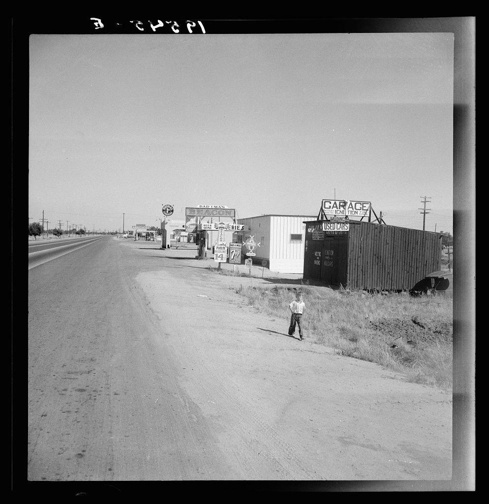 This black and white photograph depicts a desolate roadside scene with an old-style gas station on the left, featuring signs for "Dad & Son's" garage and a small sign that appears to advertise something related to food or drink. To the right stands another building labeled as a used car garage, its structure suggesting it might be made of corrugated metal.

A young boy is seen standing in the middle foreground on what looks like an old road with tire tracks visible. His posture suggests he may have been walking and stopped for a moment. The sky above is clear, indicating good weather conditions at the time of capture. Power lines run parallel to the highway, disappearing into the distance.

The overall impression given by this image evokes a sense of isolation or perhaps nostalgia, as it captures a fleeting moment in what seems like an earlier era based on its monochrome aesthetic and architectural styles.