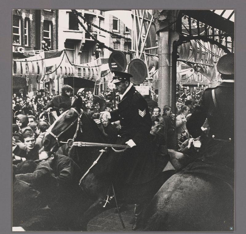 The image depicts a dynamic street scene from what appears to be the mid-1960s. A mounted police officer is in focus, wearing a formal uniform with distinguishable badges and insignia on his sleeves, which could indicate rank or unit identification. He holds onto reins tightly as he rides through the crowded streets. The crowd consists of various individuals who appear engaged in what might be a protest or public demonstration. Some people are seen reaching out towards the officer, possibly trying to communicate with him or convey their grievances.

The setting is an urban environment, likely Amsterdam's famous Raadhuisstraat (City Hall Street), as suggested by the context provided. The architecture includes buildings typical of European cityscapes from that era, characterized by narrow streets and multi-storied structures. There are signs visible in Dutch, including a "Stop verblijf" sign indicating restrictions on short-term accommodation or residency.

The black-and-white photograph captures raw emotions and tensions between the police officer and the crowd. The contrast of light and shadow adds to the dramatic effect of the scene, emphasizing the seriousness of the moment. Overall, this image portrays a historical snapshot of civil unrest during an important public event in Dutch history.