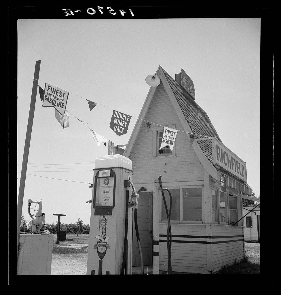 The image is a black-and-white photograph showing an old-fashioned gas station. The structure of the building has a gabled roof with decorative trim and siding, giving it a quaint appearance typical for establishments from past decades.

In front of the building stands a vintage gasoline pump labeled "Finest Gasoline" by Richfield Oil Company. Above the fuel dispenser are banners that read "Finest on Premium Gasoline," "Double Money Back," indicating promotional messages intended to attract customers with promises of better quality and refunds if not satisfied. Another banner, which reads "Double Money back or Richfield," is attached near a window.

Various other signs adorn the building's facade including one at its peak that says "Richfield" in bold letters against an ornate background featuring stars, enhancing its classic look.

Two gas pumps are visible to the left side of the image with additional fueling equipment around them. The surrounding environment appears dry and dusty, indicative of a rural or semi-desert area, which is consistent with what one might expect along highways through regions like Tulare and Fresno in California as suggested by the caption provided.