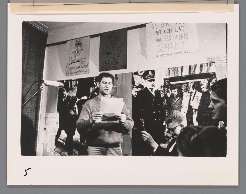 The image is a black and white photograph featuring several individuals in what appears to be an indoor setting, possibly during a public event or gathering. In the foreground, there's a man standing with his eyes closed, holding some papers which he seems to be reading from attentively. He wears a knitted sweater and has curly hair that falls onto his forehead.

Behind him, various people are visible in different states of focus; some appear engaged while others look more passive or contemplative. Among these individuals is another man dressed in formal attire resembling a police officer's uniform with badges and buttons on the chest area. He stands out prominently due to his authoritative stance and dark-colored coat. There's also text displayed above him, possibly as part of an exhibition setup.

On display around this central figure are cutouts or prints depicting different characters; one character is labeled "MEISJES," which may be a reference to the comic book series 'Smoes,' while another shows what appears to be a police officer. The words on these cutouts seem like captions, with phrases such as "AL EEN LAT MET EN LAT VAN EN SCHIM VAN GE HAD?" and "WORDT OOM AGENT," which translate from Dutch suggesting themes of communication or authority.

Additional text is visible at the top corner but seems to be partially obscured. The overall atmosphere suggests a protest [...]