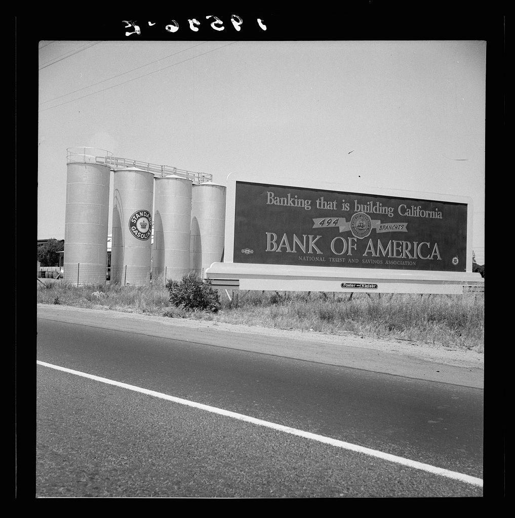 The image depicts a black-and-white photograph of an outdoor scene, presumably taken from the perspective of someone standing on or near a road. In the foreground, there's a portion of a paved road with visible white lane markings.

On either side of this roadway are large cylindrical storage tanks, likely used for storing liquids such as oil or chemicals, based on their design and industrial look. The left tank has "Standard Gasoline" labeled on it in a circular emblem, indicating the contents or brand associated with that structure.

A prominent feature is an advertisement billboard alongside these tanks. It reads: "Banking that is building California BANK OF AMERICA NATIONAL TRUST AND SAVINGS ASSOCIATION 494 BRANCHES," suggesting this image was taken at a time when Bank of America had significant branch expansion in the state, possibly during its growth phase or an advertisement campaign.

The billboard also includes smaller text and logos which are less legible due to distance from the camera. The overall condition suggests it is not modern as there's some wear on the edges of the image indicating age.