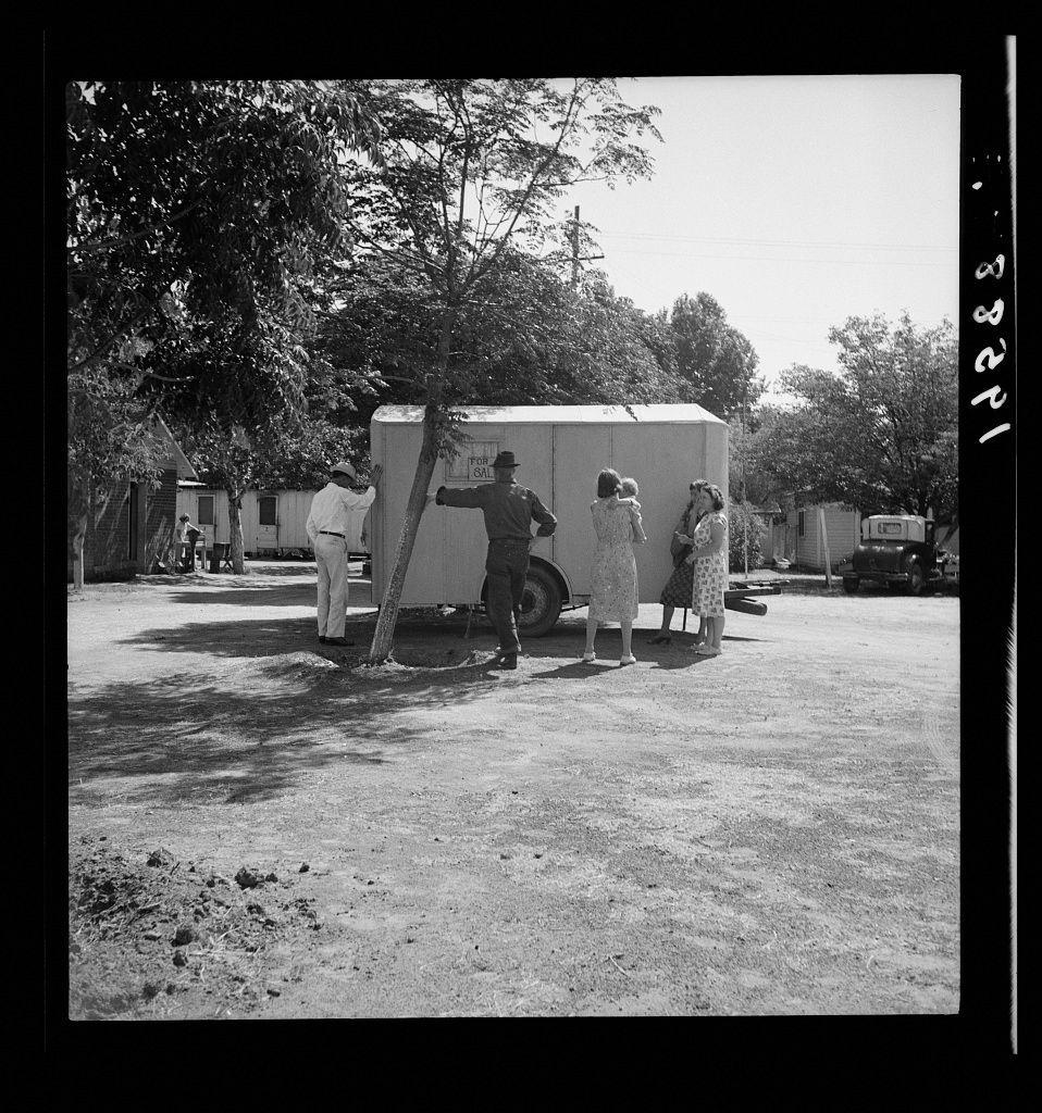 The image depicts a black and white photograph capturing an outdoor scene involving five individuals, with one of them operating a small house trailer. The setting appears to be in a rural or semi-urban area surrounded by trees and other structures.

There are four people standing near the house trailer: two women dressed in long dresses, one wearing glasses; another woman bending slightly as if interacting with someone inside the trailer; and an older man wearing a hat looking on from behind. To their right is a young person who seems to be reaching into or examining something within the trailer.

The scene suggests that they might have stopped for refreshments at this location while traveling, possibly along U.S. Route 99 between Tulare and Fresno in California during the era depicted by Dorothea Lange's work. The individuals appear engaged with their surroundings, indicating a moment of interaction or consideration regarding the trailer.

The atmosphere is casual yet somewhat curious, as if they are assessing potential for purchase or just passing through on a journey.