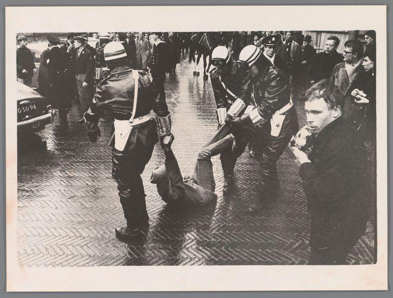 The image depicts a tense and chaotic scene on a wet pavement, likely during an urban protest or riot. Several individuals in what appear to be police uniforms are engaged with another person who is lying prone on the ground. The officers wear dark jackets, helmets, and one has visible insignia that suggests they might represent a mounted law enforcement unit.

Around them, numerous bystanders observe or react to the unfolding events. Some of these observers seem concerned or surprised, while others appear more detached. A police vehicle with 'G-5096' on its side is parked in the background, adding context that this scene may be near an official location such as a law enforcement office.

The black and white composition lends the photograph a stark, dramatic quality, emphasizing contrasts between light and shadow. The image captures a moment of physical confrontation with significant social implications, suggesting it could have been taken during a historical event or public demonstration in Amsterdam on May 6th, 1966.