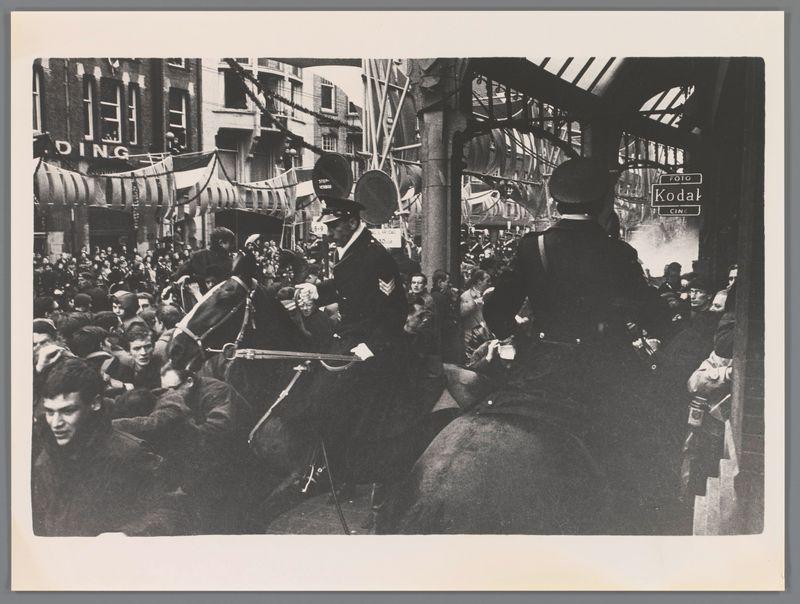 The image captures a bustling urban street scene with crowds gathered, suggesting some form of public gathering or protest. In the foreground and central focus is an officer on horseback from what appears to be a historical period judging by their uniform style, which includes a top hat and long coat indicative of early 20th-century attire. The officer seems engaged in direct interaction with individuals at street level, possibly addressing concerns or enforcing order.

Behind this figure are numerous civilians, many looking upwards towards the mounted police officers, suggesting that these figures hold some authority over the crowd below. Some citizens appear to be holding banners or placards, hinting at a demonstration of protest or public expression. The expressions on people's faces range from concern and agitation to curiosity and confrontation.

The environment is characterized by tall buildings with shop signs such as "DING," which may reference various establishments like cinemas (cine), and others that are less distinguishable due to the angle and quality of the image. Windows, awnings, and street-level activity suggest a commercial district. The photo's black-and-white nature adds an historical dimension.

The composition is dynamic with lines converging towards the center where the officer on horseback stands out as the main subject. Shadows cast by buildings i [...]
