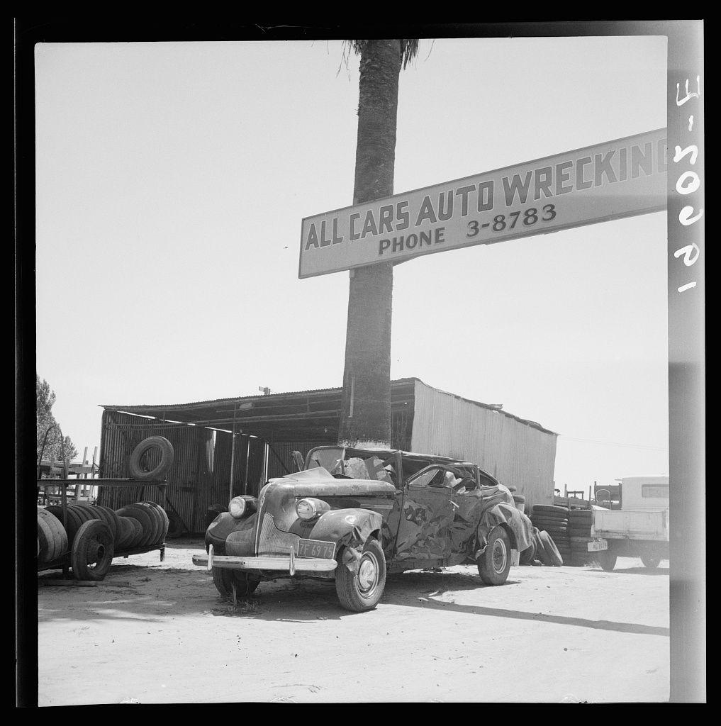 The image is a black-and-white photograph depicting an old, damaged vehicle with visible structural deformities. It's parked near what appears to be a sign that reads "ALL CARS AUTO WRECKING" and provides contact information for the establishment.

In the background, there are large oil barrels stacked horizontally on one another and vertical stacks of tires alongside some industrial structures or buildings covered in corrugated metal sheeting. The vehicle itself has its front right side crumpled and appears to have sustained severe damage overall. Its license plate is visible but not legible due to resolution constraints.

The sky above is clear, suggesting a sunny day without any clouds obstructing the sunlight. There are no people or other moving objects captured in this scene; it's focused on the vehicle and its immediate surroundings which includes the wreckage yard sign and barrels of oil.

This image captures an atmosphere that might evoke thoughts about automobile history, recycling, industrial use of space for junked vehicles, or perhaps environmental concerns regarding waste management.