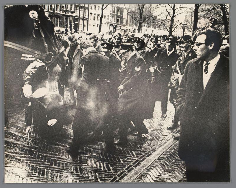 The image depicts a group of people gathered on an urban street. In the foreground, there is a horse with its head down near what appears to be two individuals crouched close together; their faces are obscured by darkness and smoke or dust in the air around them.

Surrounding this scene, various groups can be seen standing at different distances from the camera's perspective. They consist of men wearing suits, hats, and some with sunglasses reflecting light, suggesting a sunny day despite the apparent disarray on the ground. The attire suggests that these individuals are likely civilians or possibly law enforcement officers.

In the background, buildings line the street indicating an urban setting. A few trees can be spotted as well, partially obscured by smoke or dust in the atmosphere. Some people appear to be looking towards a focal point off-camera, while others engage with each other through conversation.

Overall, the image conveys tension and disarray within this public space, possibly during a demonstration or event that has disrupted normal activities on the street.