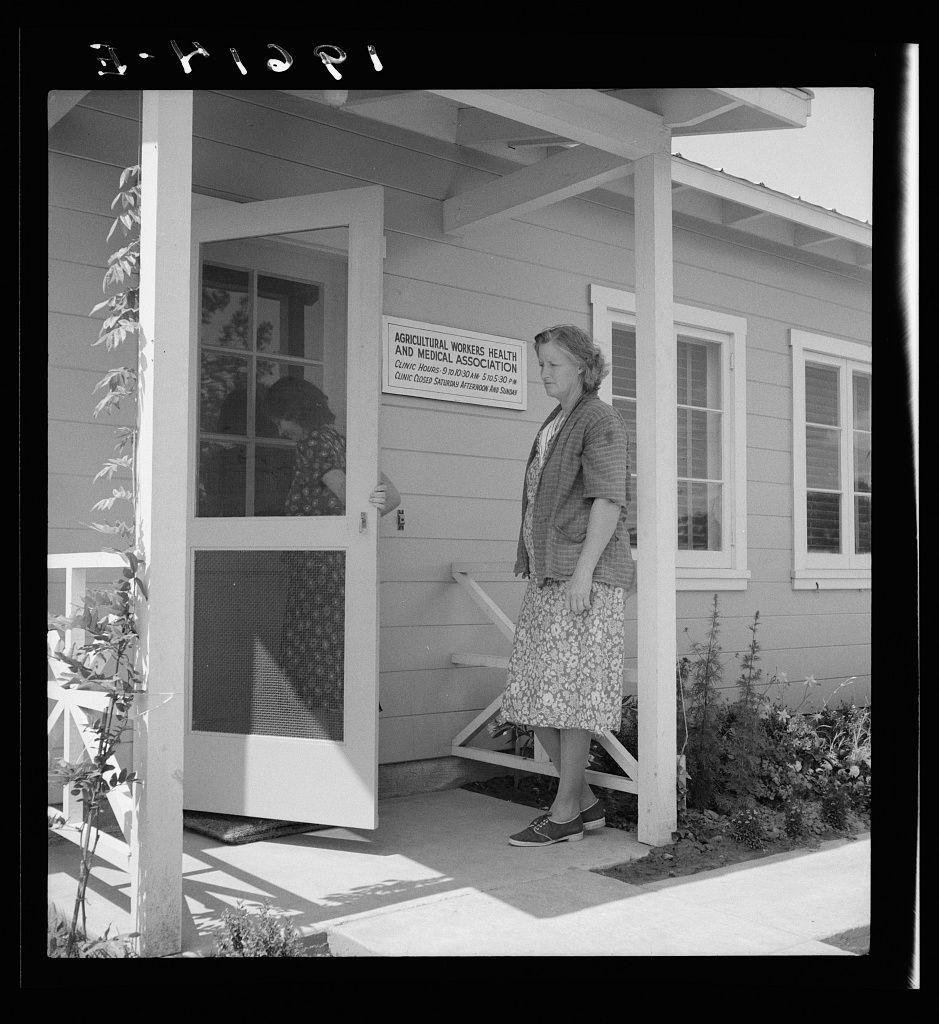 The image shows an older woman standing on the porch of a building, which appears to be a clinic. She is wearing a patterned skirt and cardigan over a blouse, with loafers as footwear. The porch has a white railing with vertical slats, and there are plants visible near the ground level alongside the structure.

A sign above the door indicates that it belongs to an association for agricultural workers' health and medical services in Tulare County, Farmersville, California. It states operating hours from 5:30 pm on Monday through Friday until 7:15 am on Saturday. The woman is looking towards someone inside or outside of the building.

The photograph has a grainy texture with high contrast between light and dark areas, typical for black-and-white images taken during historical periods.