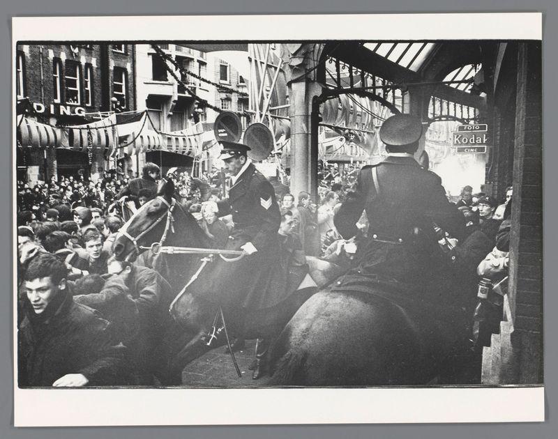 The image depicts a scene of tension or confrontation in an urban street setting, rendered in black and white. It captures several police officers on horseback amid a crowd that appears to be engaged in some form of protest or demonstration. The horses are rearing up slightly as the mounted policemen exert control over their mounts amidst chaos.

Onlookers can be seen around the scene, with expressions ranging from shock to aggression towards both the officers and each other. Various signs hang above what looks like a market area labeled 'DING', possibly indicating a specific commercial district or landmark within Amsterdam. The environment suggests it is an open-air marketplace bustling with activity before a significant event.

The photograph includes text overlays such as "FOTO" on the top right corner, identifying it likely from Roko's work during Queen Beatrix and Prince Claus' wedding in 1966 at Dam Square or possibly Raadhuisstraat. The photographer is Ed van der Elsken, a renowned Dutch street photographer known for documenting significant events through his lens.

The image conveys an intense atmosphere of public unrest or celebration with historical significance to the Netherlands during that era.