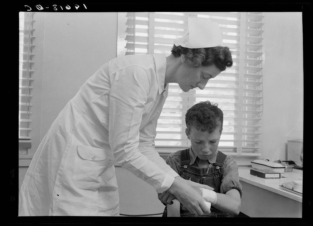 This black-and-white photograph depicts a scene where an adult nurse, dressed in a white medical uniform and cap, is assisting a young boy with what appears to be first aid. The woman holds the boy's arm steady while she applies or adjusts something around his elbow area.

The setting seems to be indoors, likely within a clinic or healthcare facility given the presence of books and papers on the table suggesting an office environment, as well as blinds covering windows in the background which provides natural light into the room. The attire of both individuals is indicative of mid-20th century fashion with distinct styles for men and women.

There are no discernible colors due to it being a black-and-white image but we can infer from the context that this was taken during or shortly after World War II era, based on clothing style and overall aesthetic. The atmosphere seems calm yet professional as the young patient appears to be receiving attentive care.