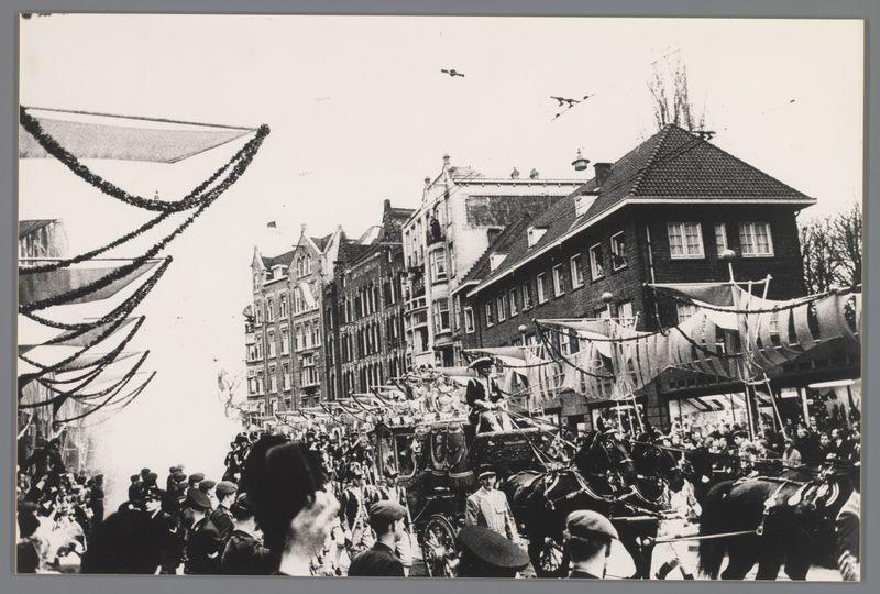 The image is a black and white photograph depicting an outdoor scene, likely from the 19th or early 20th century. In the foreground, there's a large crowd of people gathered on what appears to be a street during a public celebration or event. Amongst them are men wearing hats indicative of styles popular in that era.

In the middle ground, we see horse-drawn carriages and individuals dressed in period costumes; one carriage has figures who seem like they could be part of a ceremonial procession with swords, indicating some sort of parade or official ceremony. There's also a person seated atop what looks to be a large animal statue on wheels, suggesting it might be part of the event.

The background features several buildings that display European architectural styles, particularly those found in cities such as Amsterdam during this time period. The building facades are adorned with decorative elements like gables and ornate windows, which add to the festive atmosphere.

Adding dynamic movement to the scene is a large banner or flag being hoisted into the air by individuals on scaffolding at the top left corner of the image; it creates an impression of excitement and celebration. Above this bustling street life, birds are captured in flight against the sky, adding further vibrancy to the composition. The overall mood conveyed through these details is one of a lively public [...]