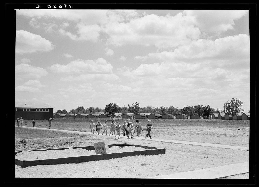 The image is a black and white photograph depicting an outdoor scene. A group of children, possibly around ages 5-8, are seen engaging in what appears to be playtime or physical activity on a dirt field with scattered patches of grass. The sky above them is partly cloudy but predominantly blue, suggesting fair weather conditions.

In the background, there's a row of prefabricated steel shelters that serve as housing for migrant agricultural workers and their families, part of a larger camp facility in Tulare County, California. These structures are uniform in appearance with pitched roofs and have multiple sections or rooms visible through open doors and windows. A few adults can be seen near these shelters.

The overall setting indicates the photograph was taken during a period when such camps were prevalent for migrant laborers who moved from one agricultural area to another following harvest cycles, requiring temporary accommodation facilities like these prefabricated steel structures. The image captures an aspect of daily life in those times and conditions, with children engaging outside while their families live nearby.

This scene is reminiscent of work done by Dorothea Lange, a photographer known for documenting the lives of migrant workers during the Great Depression era through her lens. Her photographs often provided social commentary on American society's treat [...]