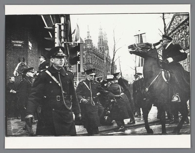 The image depicts a historical street scene captured in black and white, likely from the mid-20th century. It features several uniformed individuals on horseback and others standing or walking alongside them. The uniforms suggest they could be police officers or guards of some sort. In the background stands an ornate Gothic-style building with spires reaching into a cloudy sky. On the left side, there's signage indicating "Paleisstraat Centrum," which suggests that this scene is set in a city environment, possibly within sight of The Hague Palace given its name.

The composition captures movement and activity, as if during an event or regular patrol duty. There are no discernible modern elements like cars or street lamps with contemporary designs, supporting the historical context. Notable features include the formal attire, horse-drawn setting, and urban architecture that evoke a sense of past civic life in Europe.

Additional information about this image can be found at the provided link: Rellen bij het huwelijk van prinses Beatrix en prins Claus, Nieuwezijds Voorburgwal, Amsterdam. The photo is credited to Ed van der Elsken and dates from 1966. This indicates that it captures a moment in time during Queen Beatrix's wedding celebration on the mentioned street in Amsterdam.