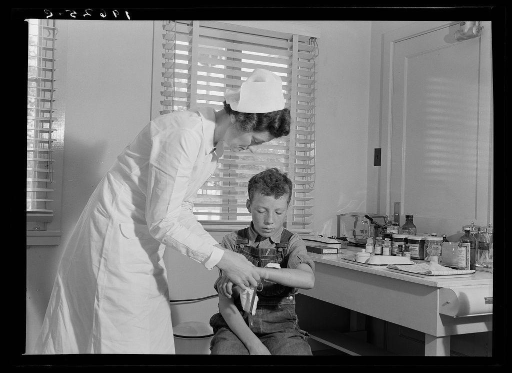 The image is a black-and-white photograph depicting an indoor scene, likely from the early to mid-20th century. A woman dressed in what appears to be medical attire - including a white coat and cap indicative of nursing or medical practice during that time period - is attending to a young boy seated on a chair beside her. The setting seems clinical, possibly within a makeshift health clinic provided by an agricultural workers' organization.

The nurse is holding out a small bottle towards the child, who appears engaged with it but not looking at the woman directly. On the table behind them are various medical and laboratory items including bottles labeled with letters or numbers, beakers, jars, containers of powders, and possibly other equipment for diagnostic tests.

There's ample natural light entering through windows fitted with blinds on both sides, illuminating a portion of the room which has white-painted walls. The environment suggests simplicity but functionality, indicative perhaps of rural healthcare settings during that era.
