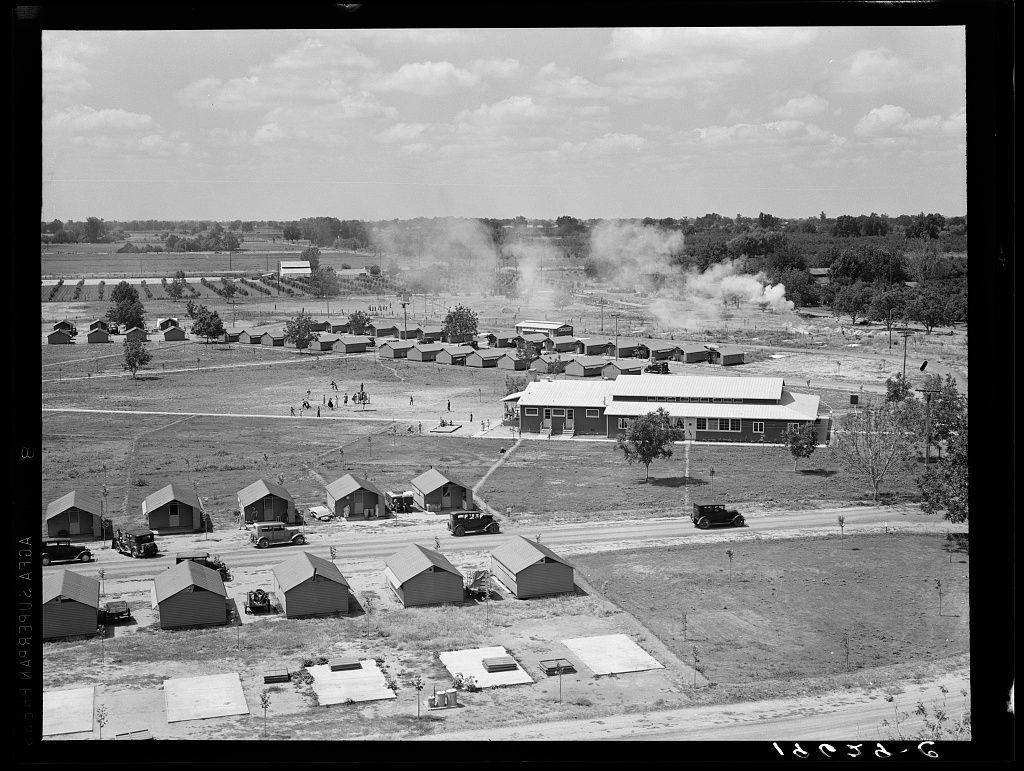 The image displays a grayscale photograph of an expansive outdoor area with numerous small structures scattered across the field. The buildings appear to be temporary or makeshift, reminiscent of military barracks but without fortifications. These are accompanied by larger community-style dwellings and what seems like a schoolhouse in view. There is visible smoke emanating from several points within this compound, suggesting active cooking or heating activity.

The setting looks rural with sparse vegetation. The sky above appears partly cloudy, indicating fair weather conditions at the time of photography. A few cars are parked on paved roads that traverse through the campsite, and a couple of individuals can be seen outside one community building. Trees sporadically dot the landscape around these structures.

In addition to this aerial perspective view of the entire layout from above what seems like an elevated vantage point—possibly a water tower or similar structure—the image contains textual data suggesting it is part of a larger collection by Dorothea Lange, possibly taken for archival purposes related to rural studies. The caption indicates that these are "Farm Security Administration (FSA) camp" likely used for housing migratory workers in Tulare County, California.

There's also a date and reference number stamped at the bottom: '1938-4'. This context implies this [...]
