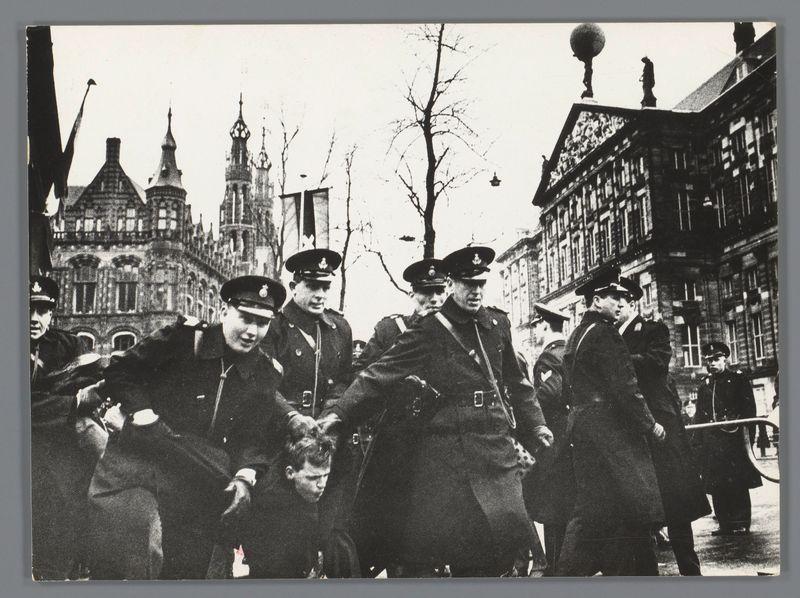 The image is a black and white photograph depicting a group of uniformed individuals, likely police officers or guards, in an urban setting. The scene appears to be from the mid-20th century based on their uniforms' style, which includes peaked caps with badges and dark coats adorned with medals and insignia.

In the foreground, there are several men standing prominently; some appear engaged in conversation while others seem vigilant. Among them is a figure who looks like he might have been apprehended or detained, as suggested by his posture being slightly bowed forward among the officers' stern expressions. His expression seems pained or resigned.

Behind this central group of figures stands another man with a raised fist and an unsmiling face; it's unclear if he has any relationship to the captured individual but suggests some level of protest or defiance in relation to the scene unfolding around him.

The backdrop features historic-looking buildings, including what appears to be Gothic architecture indicative of European cities. The trees are barren, suggesting winter or early spring months without leaves on the branches. Overhead is an overcast sky with clouds hinting at a gloomy atmosphere that might reflect the gravity of events captured in this image.

There's no explicit indication as to why these individuals have gathered; however, given their attire and demeano [...]