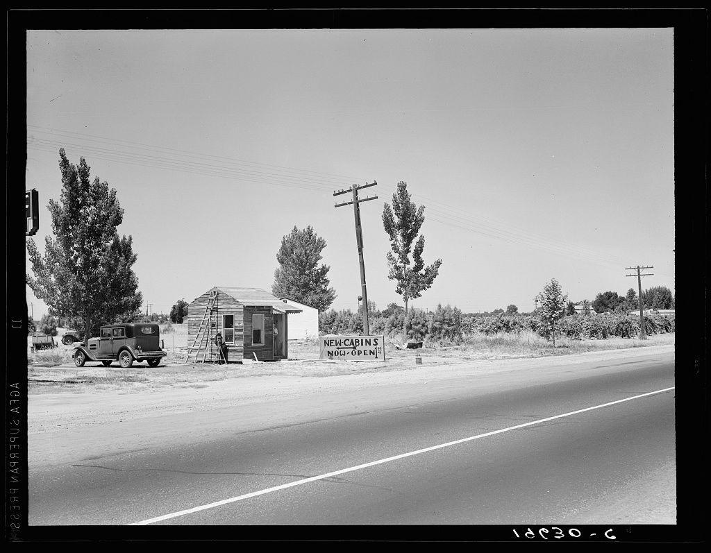 This black-and-white photograph shows a desolate roadside scene with two telephone poles, an old Model T Ford truck parked in front of a small shack labeled "NEW CABINS NOW OPEN," and another vehicle partially visible on the left. The sky is clear without any clouds, suggesting it's taken during fair weather conditions. There are trees scattered around the area which appears to be rural or semi-rural with no immediate signs of residential development close by. A road runs parallel to this scene indicating that there’s vehicular traffic passing through here.