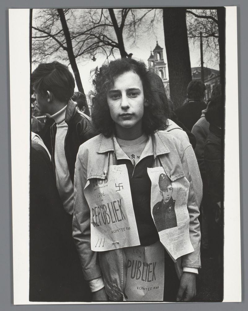 The image is a black and white photograph depicting an individual standing amidst what appears to be a crowd. The person in focus has medium-length curly hair, wears a light-colored jacket over another garment with various texts printed on it including "Publiek," which translates from Dutch as 'public.' They have several papers pinned or taped onto their clothing featuring different handwritten and printed text references such as names like "Claus" and phrases that seem to be rallying cries. The surrounding environment suggests an outdoor setting, possibly a public event with bare trees in the background indicative of either autumn or winter season. It's difficult to discern specific details due to the monochrome nature but there is enough context provided by other elements within this frame such as multiple individuals wearing coats and engaging in conversation among themselves which supports the notion that it could be some sort of social gathering, potentially a protest based on the visible texts referencing public figures or groups.