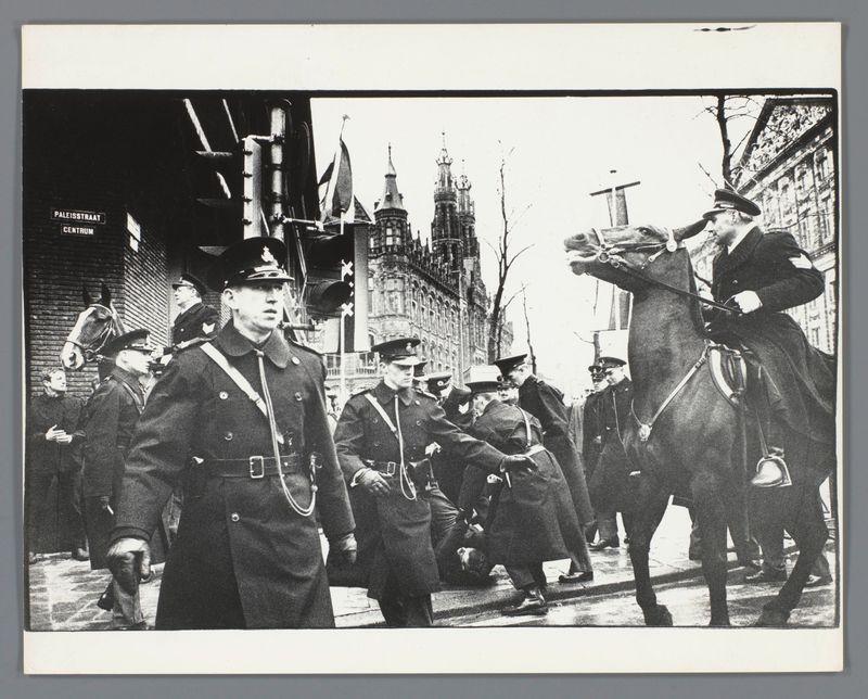 The image depicts a historical moment on Paleisstraat in Amsterdam, as indicated by the text. It shows uniformed police officers and civilians engaged in an activity that appears to be of official or ceremonial importance. The architecture suggests it's from before World War II; Gothic buildings with steeples form part of the backdrop.

One officer is mounted on a horse, directing others who are walking down the street. Their uniforms have visible insignia suggesting they're military and/or police forces. Civilians can also be seen in civilian attire but without any specific uniformity to suggest their affiliation or role within this context.

The weather seems overcast, judging by the light quality and shadows on the pavement. There is a sense of organized movement as if capturing a parade-like event or perhaps an escorting procession for someone notable off-camera.

This scene reflects historical urban life with its architectural style hinting at Europe's early 20th-century milieu.