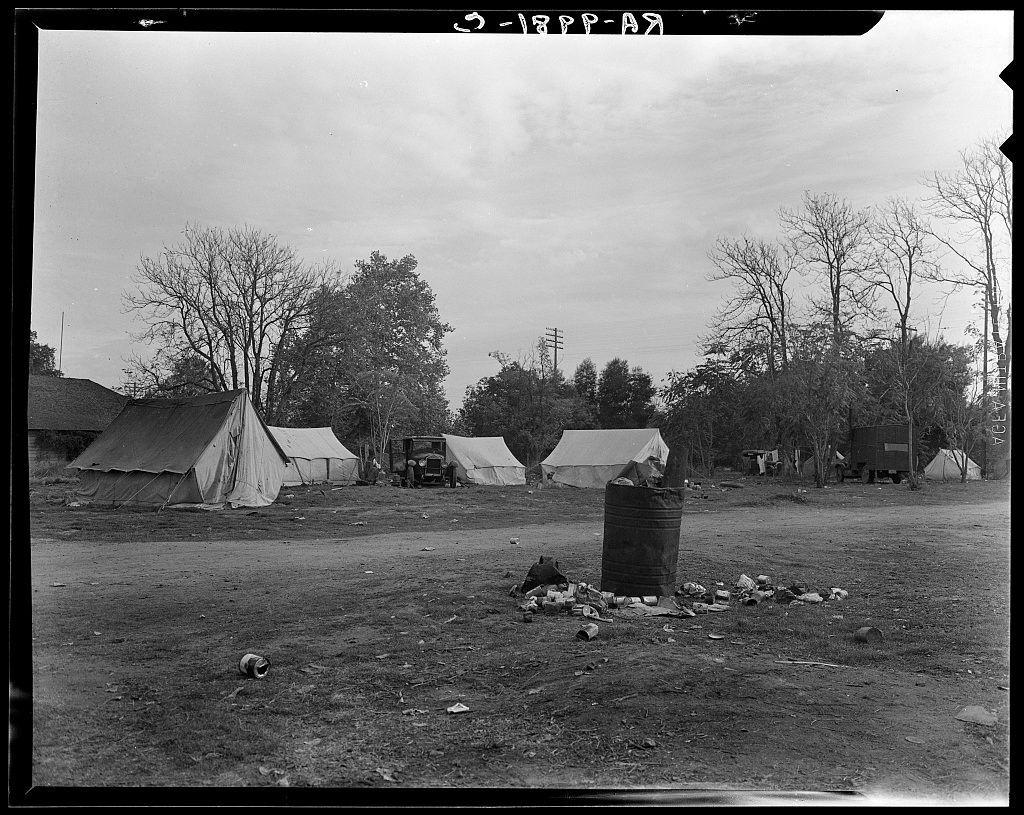 The black and white photograph captures a scene from what appears to be a temporary settlement or campsite. There are several tents pitched on the ground, suggesting that people may have been living there temporarily. The environment looks somewhat desolate with scattered debris around an overturned barrel in the foreground. In the background, trees can be seen lining the periphery of this area which could indicate it's located outside a residential zone or within a rural setting.

The photograph is marked as part of Dorothea Lange’s collection from Squatters' camp near Farmersville, Tulare County, California. The timestamp at the top right corner indicates that these images are quite old and were taken on March 1938 (March-27-45). Lange was a renowned American photographer known for her work during the Great Depression documenting social issues such as poverty, homelessness, migrant workers' struggles, industrial labor conditions, and ethnic relations.
