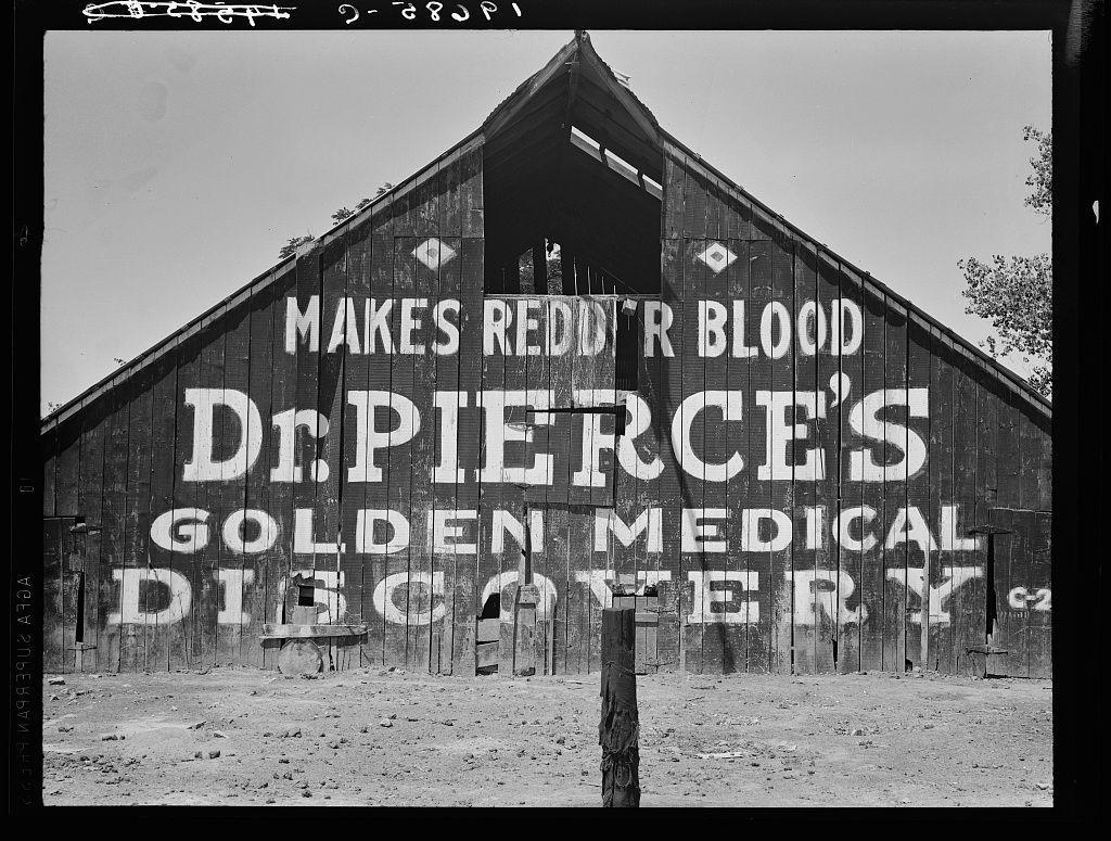 A black-and-white photograph of a large wooden barn with the words "MAKES REDDIE'S BLOOD" and other text spray-painted on it. The image has a textured, grainy quality typical of vintage photographs. It appears to be in an arid landscape based on the dry ground visible at the bottom of the frame.