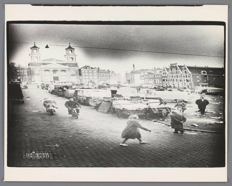 The image depicts a black and white street scene with various individuals engaging in different activities. In the foreground, there are two figures: one appears to be squatting or bending down while handling an object on the ground, possibly sorting through debris, and another seems to be walking briskly across the frame. The middle of the street is filled with scattered objects such as overturned carts, crates, and what looks like discarded furniture.

In the background stands a church-like structure with twin bell towers under a darkened sky. To its right are several buildings that appear damaged or disordered, possibly due to some form of distress or aftermath of an event. A figure on a bicycle is seen traveling across the street from left to right in this area. The overall setting has an urban decay feel, suggesting it may be taken during wartime or post-war chaos.

This photograph seems historically significant and captures a moment frozen in time that reflects social conditions possibly following World War II, given its historical context as indicated by the reference provided for Ed van der Elsken's work on Rommelmarkt.