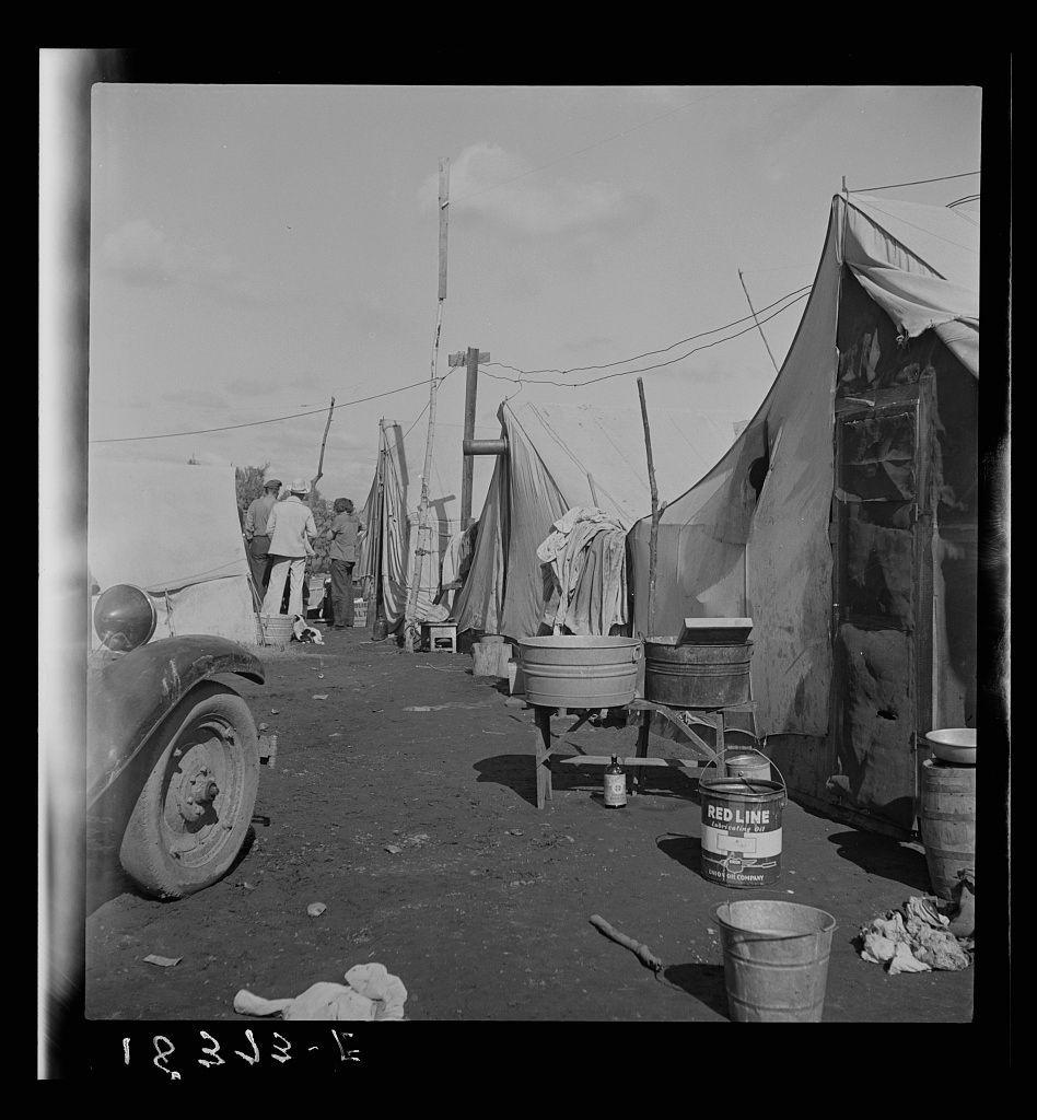 The image portrays an outdoor setting, seemingly a temporary campsite or living area. In the foreground on the left side is part of a vintage car with its wheel and fender visible. The ground appears to be dirt-covered, scattered with various objects including buckets, barrels, and what looks like laundry hanging from poles attached to makeshift shelters made of canvas tarps supported by wooden sticks.

Several people are present in the scene; one individual stands near the center of a tent-like structure while another is partially hidden behind it. The tents have open sides revealing interior details such as fold-out chairs or beds, and various personal items like clothes draped over edges. A large metal basin possibly used for washing sits under one canopy.

The sky above seems clear with no immediate signs of ongoing weather conditions affecting the scene directly. There's a sense of disarray and utilitarian living within this space which might indicate temporary housing, perhaps related to labor or travel work as suggested by references in captions mentioning "Orange pickers camp" in Farmersville, Tulare County California.

The black-and-white nature of the photograph adds an air of historical documentation with the inclusion of a date stamp at the bottom left corner reading '18.3J5-16,' likely denoting when and where it was taken or related to its archival context.
