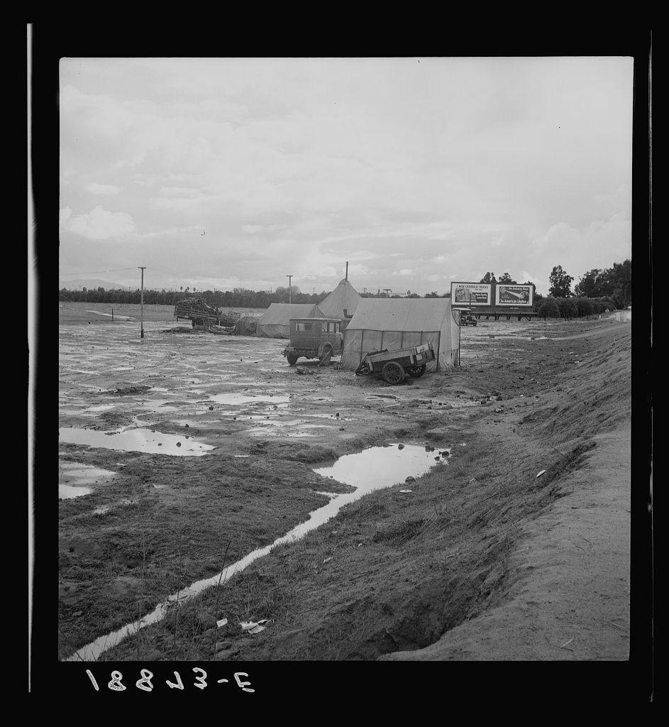 The image depicts a rural landscape with a mixture of natural and man-made elements. On the left side, there's an open field showing patches that could be crops or pastureland interspersed with bare ground. The middle section is dominated by muddy terrain with puddles reflecting parts of the scene above them. To the right lies uneven, cleared land which seems to have been prepared for agricultural purposes but currently unoccupied and awaiting cultivation.

Two large, temporary structures are prominent; they appear like mobile homes or storage units made from what looks like canvas material supported on a wooden frame. They're positioned off-center with one nearer than the other, suggesting an impromptu settlement rather than a permanent installation.

A road runs horizontally across the photo at the bottom edge and is flanked by these structures. Alongside this roadway are signs of recent activity: tracks in the soil leading away from or towards the homes suggest vehicles have been moving along here recently. The overall impression could be that of transient living conditions, perhaps indicative of migrant farm workers during a seasonal work period.

In the background, beyond the field and near what seems to be an open gate for entry/exit purposes off one side of the road, stands another large structure - possibly more housing or storage related infrastructure. A billboa [...]