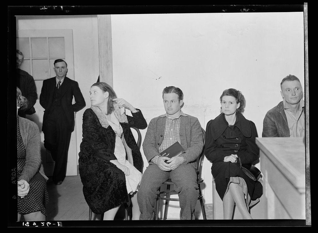 A black and white photograph depicting a group of people sitting in chairs, seemingly waiting for an important meeting or hearing. The setting appears to be indoors with plain walls, possibly within the context of government services during wartime rationing as inferred from their somber expressions and formal attire typical of that era. Some individuals are holding papers or books which could signify application forms or relevant documents pertinent to a governmental process such as farm purchase loans under the Farm Tenancy Act passed by Congress in 1937, indicating economic support measures for farmers during World War II rationing conditions.