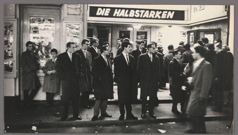 The image captures a group of men standing in front of an establishment with the sign "DIE HALBSTARKEN" prominently displayed above. The scene appears to be from mid-20th century, judging by the fashion and photographic style. Men are dressed in dark suits, coats, and overcoats indicative of formal or business attire typical for that era. Some wear ties, while others sport hats like fedoras. Their postures vary slightly; some lean forward, engaging with each other or looking towards something outside the frame.

The sidewalk is busy with people walking by, suggesting a bustling urban environment possibly in an entertainment district given proximity to what seems like a movie theater entrance labeled "Zaal" which translates from Dutch as 'hall' or cinema. The ground shows scattered litter and footsteps on concrete, typical of crowded city streets during that time period.

In the background are other storefronts with signs indicating more shops, enhancing the sense of being in an active commercial area. A woman walks by to the right side, dressed differently than the men—she wears a coat without a collar and appears less formally dressed compared to them. Her presence adds diversity to the group dynamic captured.

The image is grainy, characteristic of film photography from that period, with natural light illuminating the scene suggesting daytime or early evening hours as p [...]