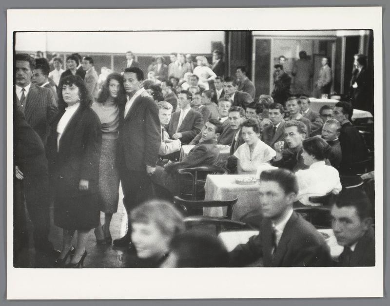 The image depicts a crowded scene where people are gathered closely together. They appear to be in various states of engagement, with some looking at the camera and others engaged in conversation or observing something off-camera. The attire is predominantly formal, suggesting that this may have been an event such as a dance hall gathering in 1950s Amsterdam. Several individuals wear hats, which was common fashion during that era. A man on the left side stands out due to his height and expressive facial expression while holding what appears to be a drink can or bottle.

The environment seems casual yet social with no visible decorations indicating a specific theme for an event such as Mardi Gras; instead it suggests a more general gathering like those held in Amsterdam's dance halls. The photograph captures the diversity of people, including individuals of varying ages and genders, adding to its historical value by showcasing societal norms from that time period.

There is no clear depiction of any objects related to Mardi Gras festivities within this image; therefore it can be concluded that such a theme was not present in what we see here.