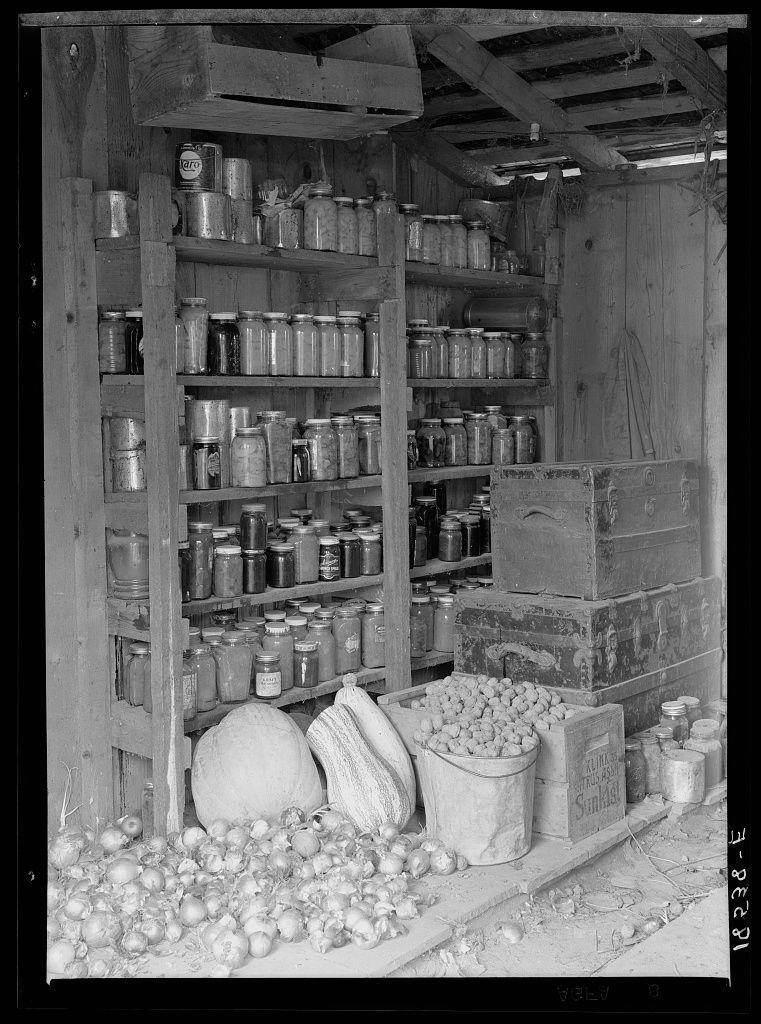 The image depicts an indoor storage area with various items. On the left side, there is a wooden shelf filled with numerous glass jars containing different substances, possibly foodstuffs or preserves. Above this shelf on the wall, more jars are arranged in rows. To the right of the shelves, several metal cans and additional jars can be seen neatly placed against the back wall.

On the floor below these storage spaces, there is an assortment of items including a large melon, what appears to be sugar cane, a bag with a white label reading "Rural Thrift Society", a box labeled "Salt," as well as numerous onions and potatoes scattered around. 

The scene has a rustic feel, indicative of rural life or early 20th-century living conditions in America. The image is black and white, adding to the vintage atmosphere. There are no people visible within this storage area, focusing solely on the stored goods.

Additional context about the photograph can be found from Rural rehabilitation documents mentioning Tulare County, California where a family had been receiving relief aid but has since established themselves through farming efforts. The cash outlay for food is reported to have reduced significantly following their re-establishment in agriculture.