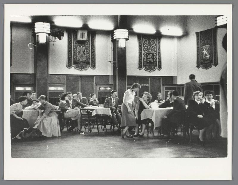 This image captures a scene inside what appears to be an old-fashioned, communal dining area. The room is filled with people seated at tables covered in white tablecloths, engaged in conversation or enjoying their meals. In the foreground, a waitress dressed in period-appropriate attire serves customers. She moves through the crowd, attending to various groups of diners who are either conversing animatedly among themselves or focused on their individual dining companions.

The walls above the tables feature ornate banners with intricate designs and inscriptions that suggest a formal or semi-formal establishment. The lighting is provided by large pendant lamps hanging from the ceiling, casting an even glow across the room. In the background, more people can be seen, some standing near the entrance while others are seated at additional dining areas.

The overall atmosphere suggests a busy social setting, possibly in Amsterdam's Nieuwendijk area during the mid-20th century, as indicated by the provided context from Ed van der Elsken. The composition of this photograph emphasizes the bustling environment and captures a moment frozen in time within an era that values communal dining experiences.

For additional information about this image, please refer to Jongeren in een danszaal op de Nieuwendijk (Young people at a dance hall on the Nieuwendijk) by Ed van der Elsken from 1955.