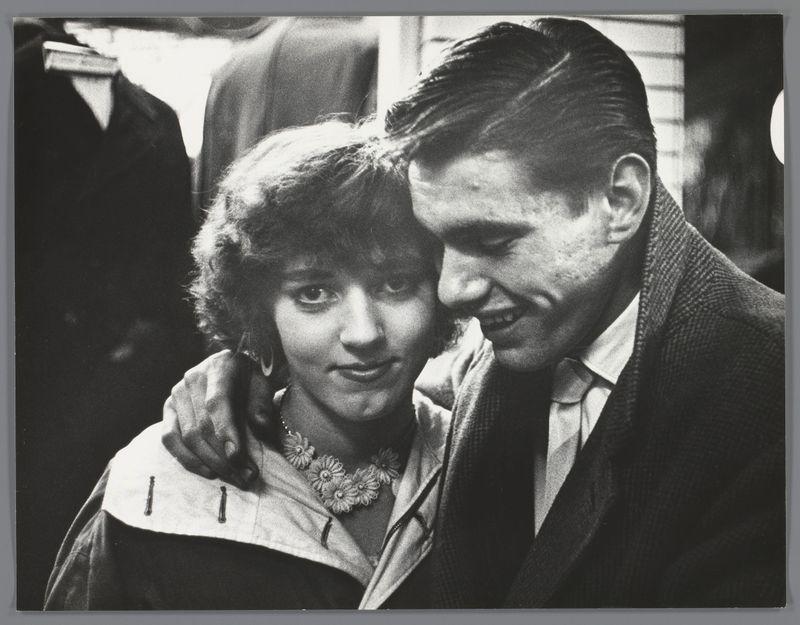 This black and white photograph depicts a man affectionately leaning his head against the shoulder of another person, who is partially turned away from the camera. The background suggests an urban setting with structures that could be buildings or street elements. Both individuals are dressed in what appears to be period-specific attire; the man wears a suit and tie, while the other has on a lighter garment adorned with flowers at the collar area, possibly indicating a floral accessory like lace or embroidery. There's no discernible text within this image aside from potential context clues related to its historical setting.

To provide additional information about this photograph: it is likely part of Ed van der Elsken’s photographic series documenting youth culture in Amsterdam during 1956. The individuals captured could be participants at a social event or simply people encountered on the street by the photographer, who was known for his candid and intimate approach to capturing life moments with spontaneity.

The photograph's historical significance is further underscored by its medium of black and white film photography, which served as an early form of mass communication before color photographs became widespread. Ed van der Elsken’s work often portrayed a sense of authenticity and connectedness that stood out in his documentation of urban life during the mid-20th ce [...]