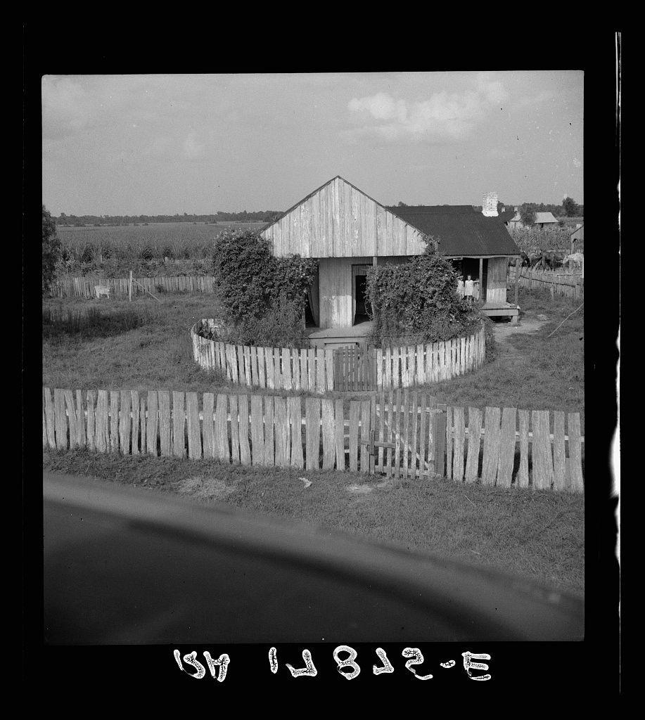 The image is a black and white photograph depicting an old wooden cabin with a fenced enclosure. The structure appears rustic, featuring weathered planks and overgrown vegetation climbing its walls. A single chimney rises from the roof of what seems to be a larger adjacent building or barn. Surrounding this setup are fields indicative of farmland, suggesting a rural setting. In front is a visible fence bordering the property with more agricultural land stretching into the distance under an open sky with sparse cloud coverage. The photograph bears handwritten text at its bottom reading "518-627-3" and additional cursive that might be personal annotations or identifiers but are not fully legible due to handwriting style and size.