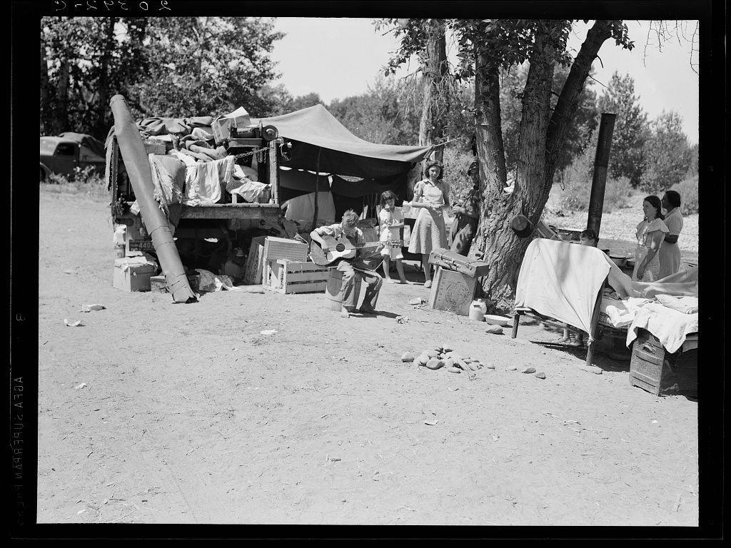 The black and white image depicts a group of people gathered around what appears to be an outdoor campsite. The setting is rustic, with several individuals seated on crates or benches under the shade provided by makeshift tents made from fabric draped over poles.

In the center foreground, there's a person sitting cross-legged playing a stringed instrument, possibly guitar. To their left, another individual reclines in what seems like relaxation while holding onto some objects that could be personal belongings. In front of them is an assortment of items strewn on the ground and atop wooden crates: clothing, books or magazines, boxes likely containing supplies, and miscellaneous other household goods.

To the right side of the image stands a woman with her back to us; she appears engaged in conversation with another person partially visible beside her. They are surrounded by more camping equipment—clothes strewn about on folding chairs, wooden crates used as makeshift tables or storage containers—and an open box possibly containing foodstuffs.

The background is filled with trees and vegetation indicative of a rural environment. A vehicle parked further back suggests that this gathering could be connected to travel for work such as migrant farm laborers during the Dust Bowl era.

This scene evokes themes of resilience, community amidst hardship, or temporary living situati [...]