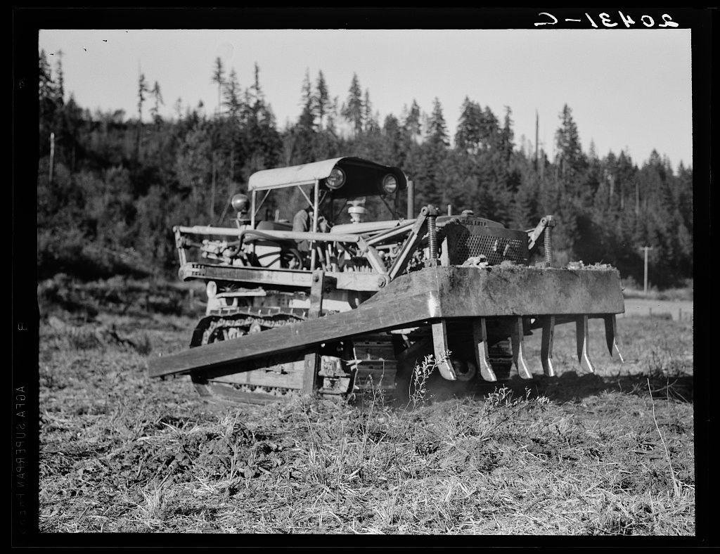The image is a black-and-white photograph depicting an old-fashioned agricultural tractor in operation. The vehicle, seemingly made from metal with wooden components, has large wheels and is equipped with a long blade at the front for tilling or plowing through the field's vegetation. It appears to be situated on uneven terrain surrounded by shrubbery and trees indicative of a forested area. The sky is clear without any visible clouds. There are no people in view within this particular frame, suggesting that the tractor may have been momentarily unattended during work hours.
