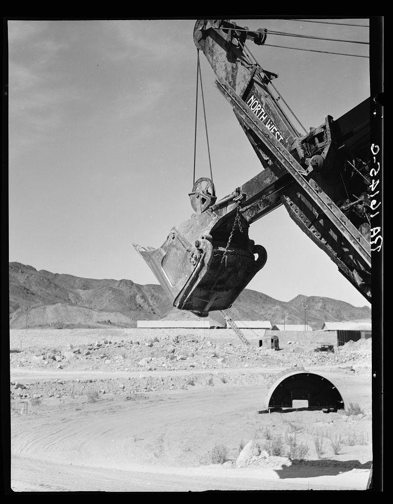 The image is a black and white photograph depicting an industrial scene. A large crane with the words "NORTH WEST" on its arm extends from the top left corner into the frame, holding what appears to be material in its bucket. The background shows barren land with sparse vegetation, mountains in the distance under a clear sky, and various structures including a tunnel entrance in the lower right side of the image.

The photograph seems historical, suggesting it may have been taken during or shortly after World War II given the reference "Laguna Dam" which could be associated with war-related infrastructure. The caption also mentions an irrigation canal called All American Canal extending desert land to cultivation by irrigation in the southwestern United States. However, there is no direct evidence of such a canal within this particular image.

The overall composition gives off a stark industrial atmosphere, emphasizing human engineering against the backdrop of nature's rugged landscape.