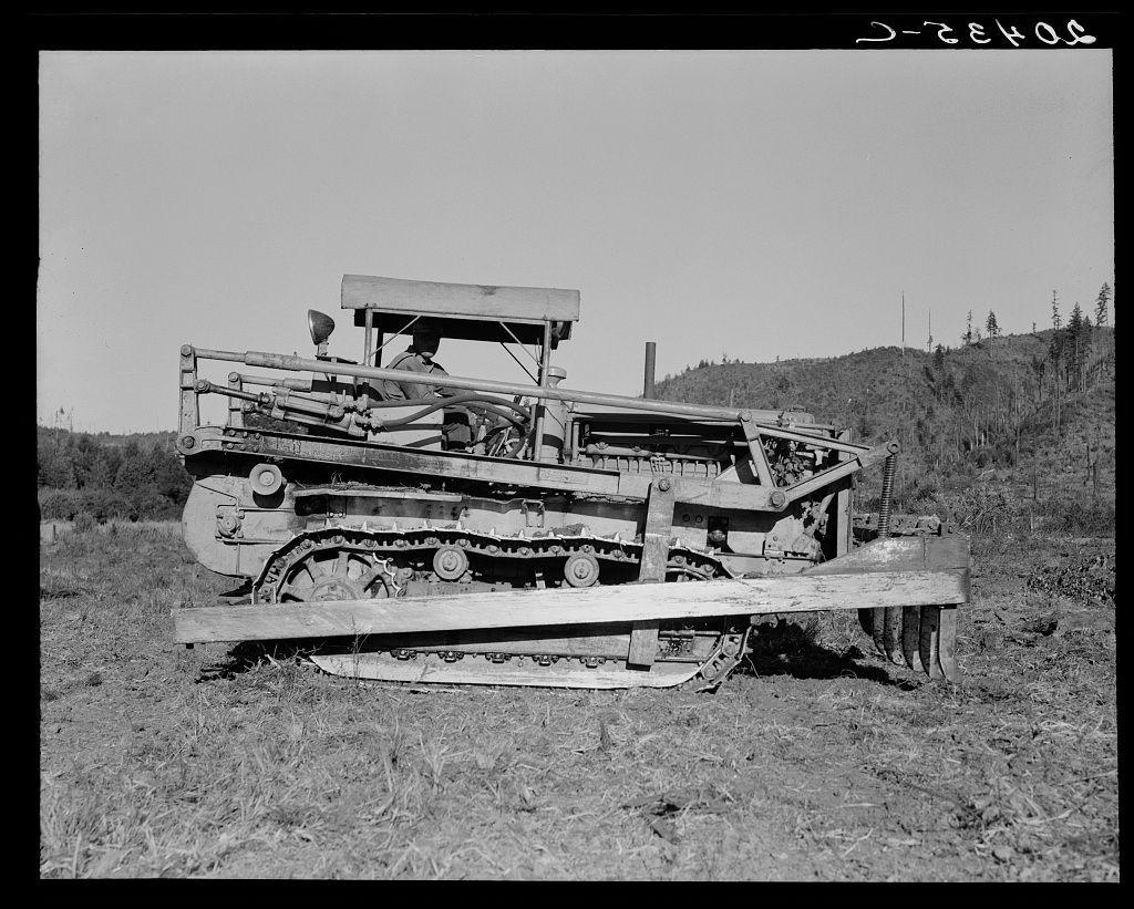 The image is a black and white photograph depicting heavy machinery used for clearing land. The machine appears to be an older model of caterpillar equipment, with large tracks on the ground suggesting it's designed for rough terrain or construction work in undeveloped areas.

It features several mechanical components such as rollers, levers, and hydraulic systems that indicate its use might involve earth-moving tasks like grading or preparing soil. The machine is parked outdoors, possibly a field given the visible natural surroundings with sparse vegetation and some trees at a distance.

There's no visible sign of modern technology, suggesting it could be from an earlier period when such equipment was more common in rural land-clearing activities. Given its context within Western Washington as mentioned in accompanying text references to Lewis County (https://images.loener.nl/DorotheaLange/full/6495/6495b7f4ba25398d90c4f8f1.jpg), this piece of equipment could be part of the historical record documenting land development or agricultural expansion in that region.