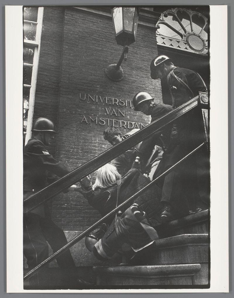 The image is a black and white photograph depicting a scene with individuals in military or police attire, along with others who appear to be civilians. They are on the steps of what seems to be an institutional building labeled "University van Amsterdam." The individuals are dressed in dark uniforms, some wearing helmets, suggesting they may be part of law enforcement or security forces dealing with a situation involving civilians. One person is being assisted up the stairs by another individual while others look on. The scene has a tense and confrontational atmosphere. There's visible text "UNIVERSITET VAN AMSTERDAM" inscribed into the brickwork above, indicating the location of this event as University van Amsterdam in 1969.