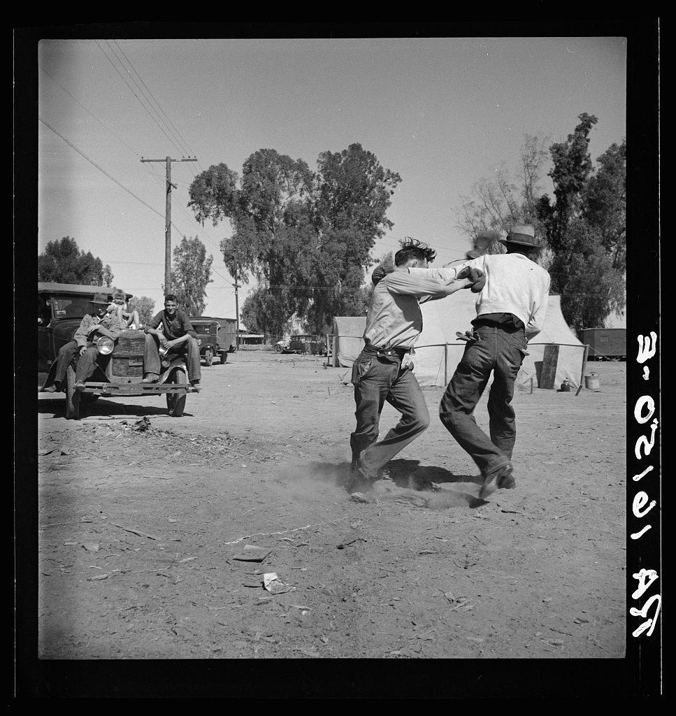 The image depicts two men engaged in a physical altercation on what appears to be a dirt road. The man wearing the light-colored shirt and dark pants is throwing punches towards another man who is dressed in a white long-sleeved shirt, jeans, and boots. This second individual seems to have been struck by one of these blows as he falls to his knees on the ground.

In the background, several individuals are gathered around an older vehicle with exposed wiring and missing parts, suggesting it may be under repair or not currently in use. The group appears focused on something off-camera, possibly observing another part of their campsite that is out of view.

Trees line both sides of the dirt road, indicating a rural setting. Tents can be seen farther back from where this altercation occurs, implying temporary housing which could suggest displacement due to migration or transient labor work conditions such as farmworkers' camps.

The black and white image's graininess indicates it may have been taken with an older camera model without modern digital processing capabilities for color correction. There is a date stamp in the lower right corner of the photo reading "Feb 18, '45", which places this moment within World War II-era America when many people were displaced by war or moving as part of the Great Migration due to economic conditions.

The scene captures a snapshot of life [...]