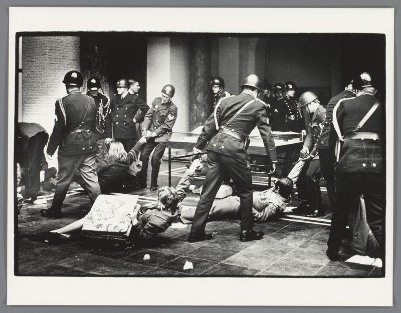 The image depicts a tense moment involving multiple individuals in what appears to be an indoor setting with tiled flooring. In the center of the action, several people are involved in a physical altercation or restraint scenario.

At least five men wearing military-style uniforms and helmets are actively restraining two younger-looking males on the floor, who appear disoriented and possibly injured. One man is kneeling over one person while another holds onto their arms, suggesting they might be trying to subdue them quickly for some reason. The expressions of shock or distress can be inferred from their facial reactions.

Around this central event are several observers, including men in similar military uniforms standing by with neutral postures and a few others observing the situation intently but without direct involvement. There is also an individual wearing casual clothing who seems to have fallen on the floor amidst the chaos.

The environment suggests it might be part of a larger public gathering or protest that has taken place, given its urban look. The overall black-and-white tone adds a dramatic and possibly historical context to the scene depicted in this image by Ed van der Elsken from 1969 as per the provided source link.