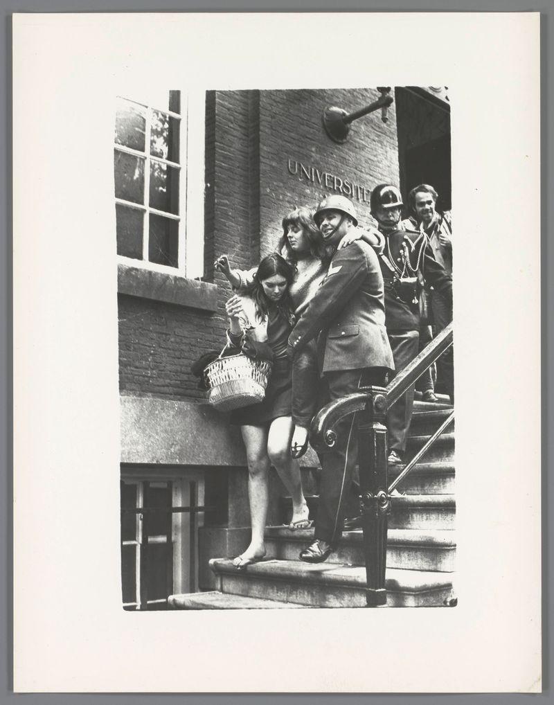 This black and white photograph captures a moment on the steps of what appears to be an educational institution, as indicated by the sign "UNIVERSITY" in capital letters engraved into the stone above. A group of individuals is descending these stairs; three are clearly visible: two women walking down side-by-side with one woman slightly ahead, holding onto her companion's shoulder for support or guidance. Both women wear short skirts and dresses, respectively—one carries a woven basket on her arm—suggesting casual attire possibly associated with leisure activities such as shopping or socializing.

At the rear of the group is another individual in what seems to be military uniform—a helmet and formal clothing that hints at a connection to armed forces; they are partially obscured by those ahead. The setting appears urban, given the architecture and style of dress common during mid-20th-century Europe.

The photograph has an air of candidness, as if it were taken in real-time without any staging or posing from its subjects—capturing everyday life with a snapshot that encapsulates social interactions and fashion norms of the era. The image is credited to Ed van der Elsken via Ontruiming van het bezette Maagdenhuis, Amsterdam [1969], as indicated by the reference in the description provided.