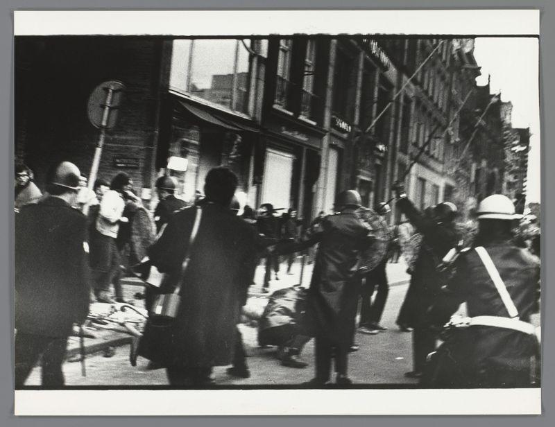The image captures a tense street scene in black and white, suggestive of an event involving conflict or protest. In the foreground, there are individuals wearing helmets, some carrying objects that resemble clubs or sticks, indicating they may be police officers equipped for crowd control. They appear to be engaging with others who seem to be protesting or fighting back against them.

Several people without protective gear engage in what appears to be a physical altercation, pushing and shoving at each other. The expressions on their faces are not clearly discernible due to the motion blur and angle of capture. Some individuals are wearing casual clothing while one person is dressed more formally with gloves that could suggest they were part of an organized group or movement.

In the background stands several rows of buildings, possibly residential apartment blocks common in urban settings from a few decades ago. The architecture suggests this might be taken during mid-20th-century Europe or North America.

The street looks narrow and crowded, hinting at high population density typical for city centers. There's a mix of urgency and chaos captured through the crowd's movements and expressions, along with the disarray caused by what seems to have been some sort of disturbance on the ground covered in debris and objects from earlier events not visible here.

There are no cl [...]