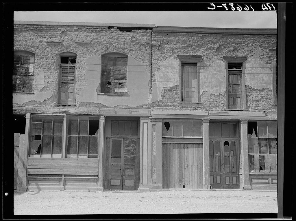 The image is a black and white photograph depicting an old building with significant signs of wear and decay. The structure appears to be made primarily from weathered stone, showing extensive cracks, missing or broken sections, and peeling plaster on the facade. Multiple windows are visible; some have boarded-up openings while others show glass panes that seem aged or damaged. There is a noticeable absence of modern elements like electrical fixtures or contemporary architectural styles.

The building has two main levels with varying window arrangements on each floor: one level features large, multi-paned windows and another displays smaller, simpler windows without any visible curtains or blinds. The ground-level section includes wooden doors, some seemingly closed while others are slightly ajar or missing panels entirely, suggesting the establishment may have once housed businesses now abandoned.

The surrounding area is not clearly depicted but seems desolate with no vegetation or other structures in close proximity to this building. This setting evokes an atmosphere of neglect and historical abandonment, which might be indicative of larger socio-economic conditions during its time period as a thriving mining town such as Tombstone, Arizona.