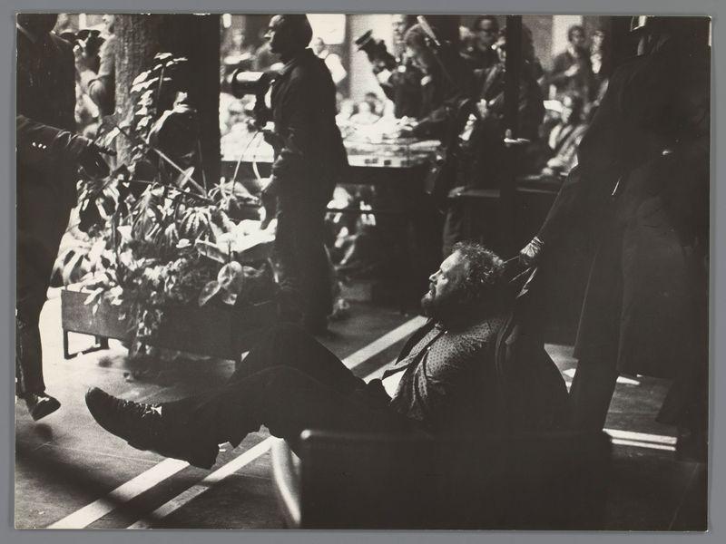The image depicts a scene with multiple individuals in an indoor setting, possibly a gallery or exhibition space. The focus is on two people; one person is reclining comfortably across the foreground floor, seemingly relaxed and contemplative, while another individual stands behind them, appearing to be engaged in conversation or interaction.

In the background, there are several other figures who seem occupied with various activities, such as examining objects or engaging with others. The environment includes potted plants that add a touch of greenery to the scene. A large plant is particularly prominent on the left side. Lighting appears natural and diffused, suggesting daytime. The overall ambiance feels dynamic yet casual.

The image captures candid moments among individuals who seem immersed in their activities within this communal space.